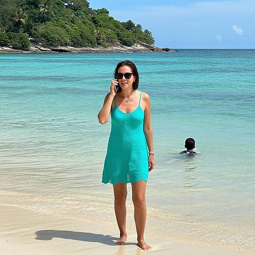 Photograph of a woman in a turquoise dress, sunglasses, and brown hair, standing on a sunny, clear sandy beach, with a person swimming in