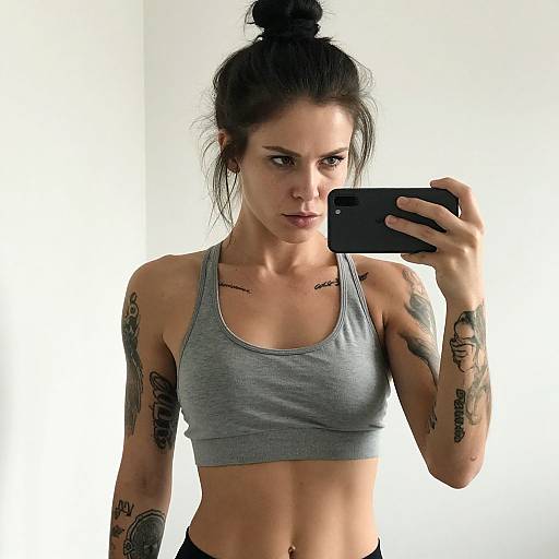 Photograph of a tattooed, dark-haired woman with a bun, wearing a gray sports bra, taking a mirror selfie with a black smartphone against a