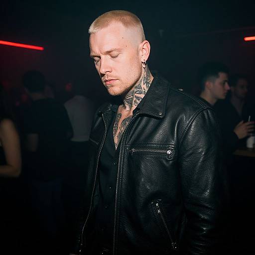 Photograph of a tattooed, bald man with fair skin, wearing a black leather jacket, in a dimly-lit, crowded nightclub. Red