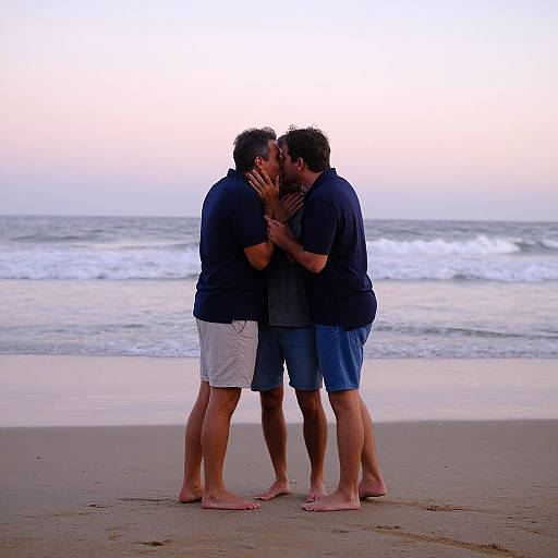 Photograph of two men kissing passionately on a sandy beach at sunset, both wearing navy shirts and shorts, standing barefoot.