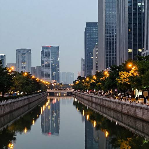 Photograph of a calm urban canal at dusk, reflecting city skyscrapers and illuminated streetlights, lined with trees and people.