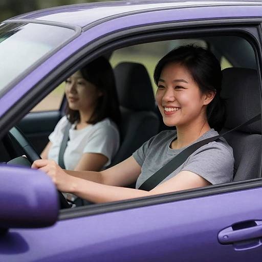 Photograph of two Asian women in a blue car; foreground woman smiles, wears gray shirt, drives, seatbelt on; background woman in white shirt
