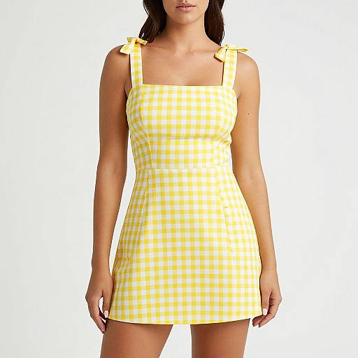 Photograph of a woman wearing a yellow and white checkered sleeveless apron dress with bow ties on straps, standing against a plain white background.