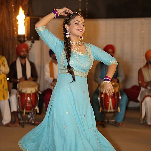 Photograph of a smiling South Asian woman in a light blue traditional dress, dancing with a braid, adorned with jewelry, in front of musicians and