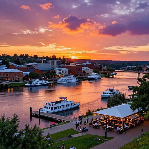 Vibrant sunset over a river with white boats, docked near a lively waterfront restaurant, surrounded by buildings and greenery. Photorealistic photograph
