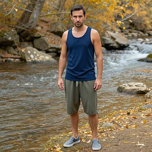 Photograph of a muscular, bearded man in a navy tank top and gray shorts, standing by a rocky autumn creek.