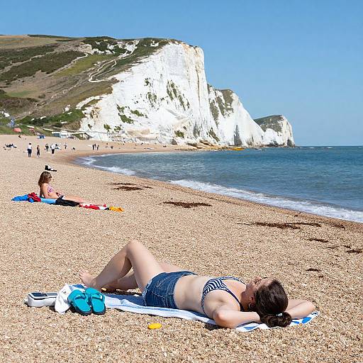 Photograph of a sunbathing woman in a striped bikini on a pebble beach, white cliffs in the background, with a clear blue sky and