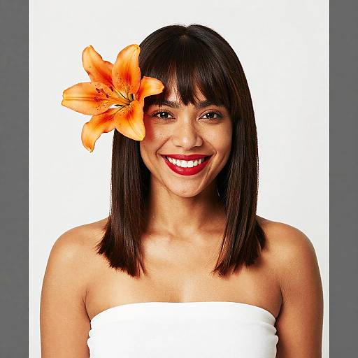 Photograph of a smiling woman with straight dark brown hair, wearing a white strapless top, and an orange lily flower in her hair, against