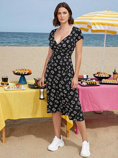 Photograph of a woman with short brown hair, wearing a black floral dress and white sneakers, standing on a beach with picnic tables covered in yellow and