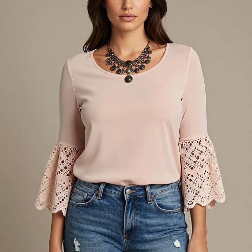Photograph of a woman with medium skin tone, wearing a pale pink blouse with lace sleeves, black statement necklace, and blue distressed jeans, standing against