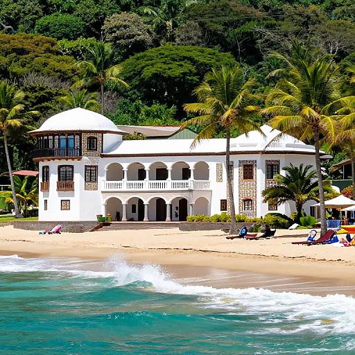 Photograph of a white, colonial-style beach house with arches and balconies, surrounded by lush green palm trees, on a sandy shore with turquoise