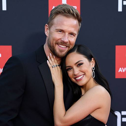 Photograph of a smiling couple at a red carpet event; man in black suit, woman in black strapless dress, dark hair, earrings. Background