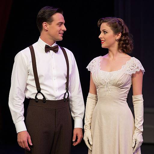 Vintage photograph of a smiling couple on stage; man in white shirt, black bow tie, suspenders; woman in white lace dress, gloves.