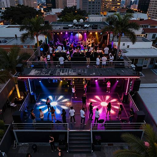 Aerial photograph of a rooftop bar at night, illuminated with colorful neon lights, surrounded by palm trees and city skyline. Crowds of people gather below