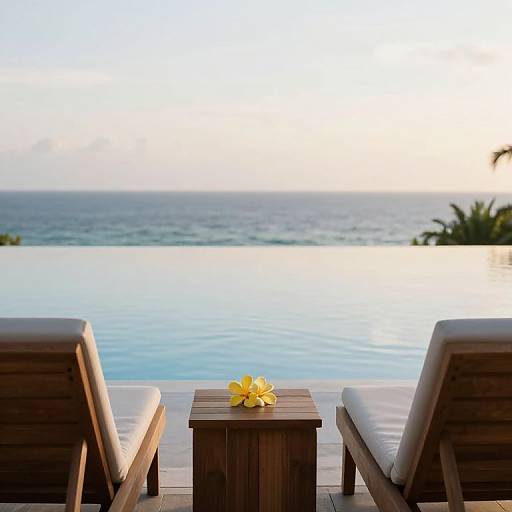 Photograph of two wooden lounge chairs with white cushions facing an infinity pool overlooking the ocean at sunset, with a yellow plumeria flower on a square