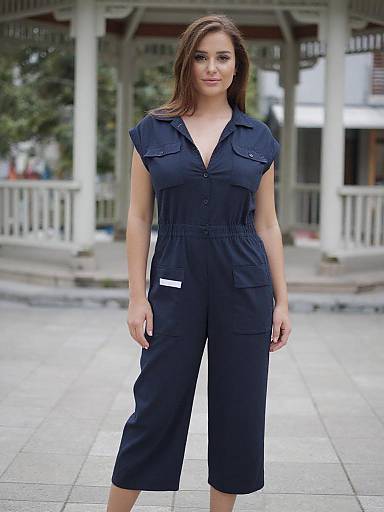 Photograph of a brown-haired woman in a navy blue, button-up, knee-length jumpsuit standing on a tiled sidewalk in front of a white,