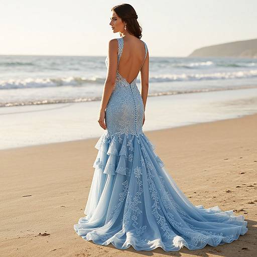 Photograph of a brunette woman in a backless, light blue, lace mermaid gown standing on a sunlit beach with waves in the background.