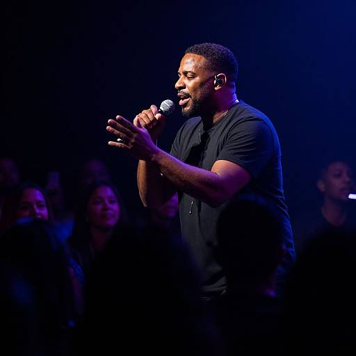 Photograph of a muscular Black man with short hair and beard, wearing a black shirt, passionately singing into a microphone on a dimly lit stage with
