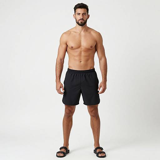 Photograph of a muscular, bearded man with medium skin tone, black shorts, and black sandals, standing against a plain white background.