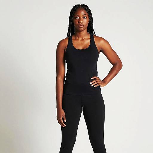 Photograph of a confident Black woman with braided hair, wearing a black tank top and pants, standing against a plain white background, with one hand
