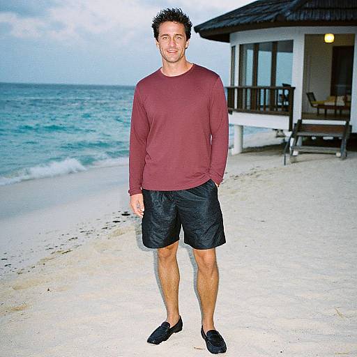 Photograph of a smiling, short-haired man in a maroon long-sleeve shirt, black shorts, and black shoes standing on a sandy beach