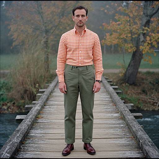 Photograph of a man with short dark hair, wearing an orange checkered shirt, green pants, and brown shoes, standing on a wooden bridge in