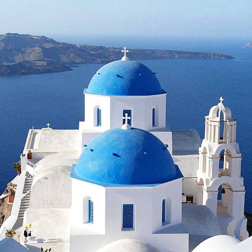 Photograph of a Greek island church with bright blue domes, white walls, and a cross-topped bell tower, overlooking a calm blue sea.