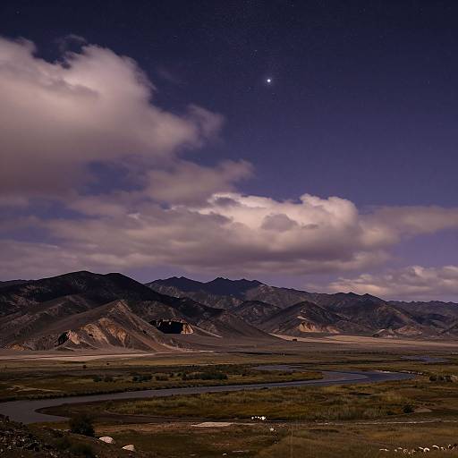 Photograph of a starry night sky over a mountainous landscape with dark, rocky peaks, a winding river, and scattered clouds. Signature in bottom