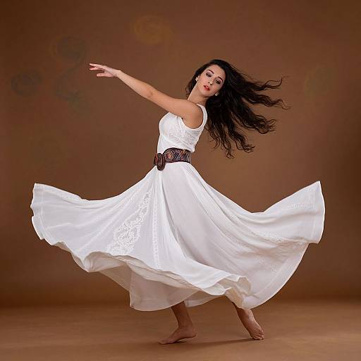 Photograph of a beautiful woman with long black hair, wearing a flowing white lace dress and brown belt, barefoot, dancing against a brown backdrop.