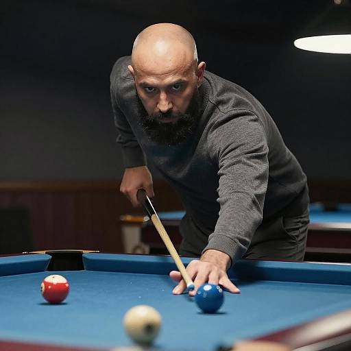 Photograph of a bald, bearded man in a black sweater, intensely aiming a pool cue at a blue ball on a blue pool table.