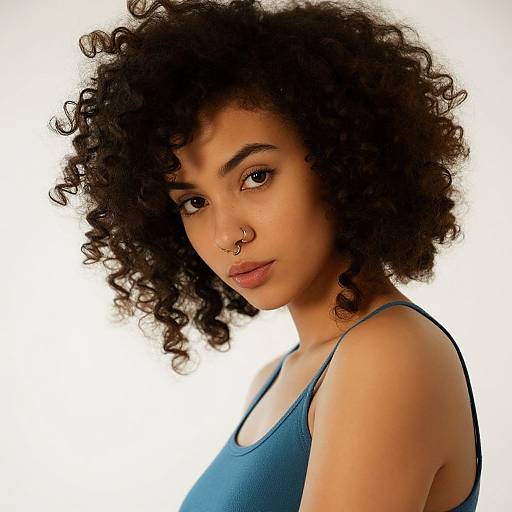 Photograph of a young woman with curly black hair, brown skin, wearing a blue tank top, nose ring, and neutral expression against a white background