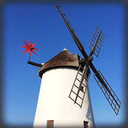 Photograph of a white, cylindrical windmill with a brown, tiled roof, black blades, and a red star-shaped wind vane against a bright