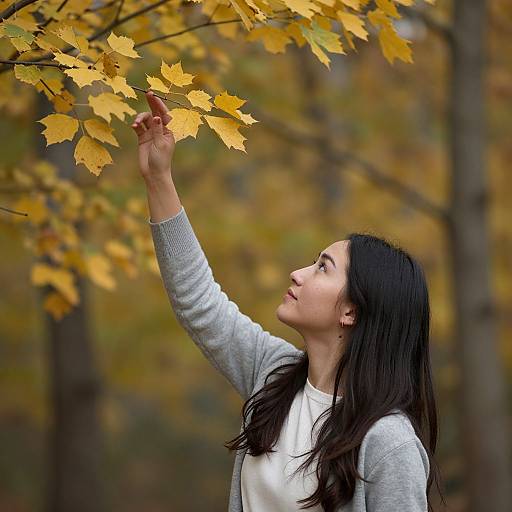 Photograph of an Asian woman with long black hair, wearing a gray cardigan and white shirt, reaching up to touch autumn yellow leaves in a forest