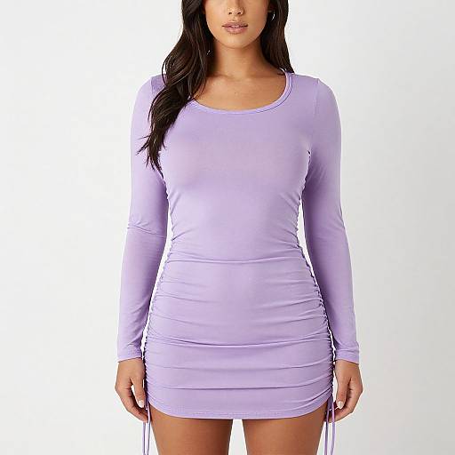 Photograph of a woman with long dark hair in a tight, long-sleeve, lavender mini dress, standing against a plain white background.