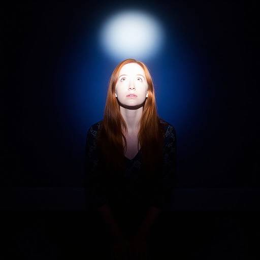 Photograph of a woman with long red hair, illuminated from above by a bright white light, against a dark blue-black background.