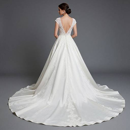 Photograph of a woman in an elegant white, backless, lace-trimmed wedding gown with a long, flowing train, standing against a gray