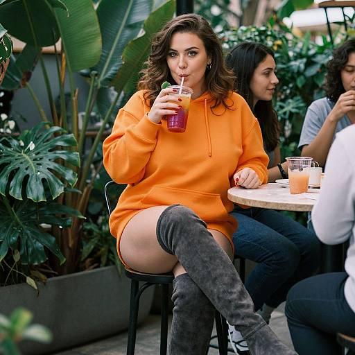 Photograph of a curly-haired woman in an orange hoodie and grey knee-high boots, sipping a purple drink outdoors, surrounded by plants and seated at