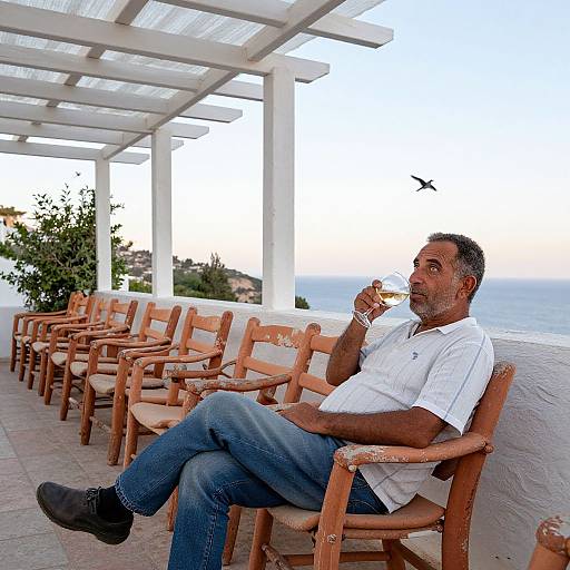 Photograph of a middle-aged man with short gray hair, wearing a white shirt and blue jeans, sipping a drink while seated on a wooden chair