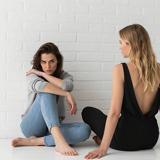 Two Women Sitting on White Floor Against Brick Wall