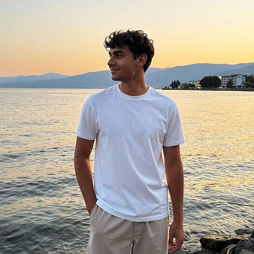 Photograph of a young man with curly brown hair, wearing a white t-shirt and beige pants, smiling at a sunset over a calm lake with mountains