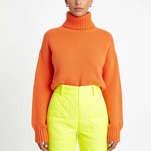 Photograph of a woman wearing an orange turtleneck sweater and high-waisted, bright yellow pants against a plain white background.