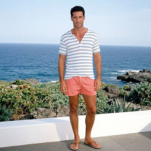 Photograph of a fit, smiling man with short dark hair, wearing a white striped shirt and coral shorts, standing on a coastal terrace with ocean and