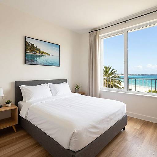 Bright, modern bedroom photograph: white bed, gray headboard, large window with ocean view, palm plant, wooden floor, framed beach photo, white