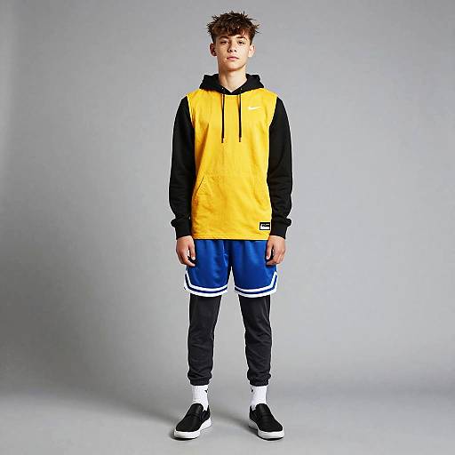 Photograph of a young boy with spiky brown hair, wearing a yellow and black hoodie, blue athletic shorts, black pants, and black sneakers,