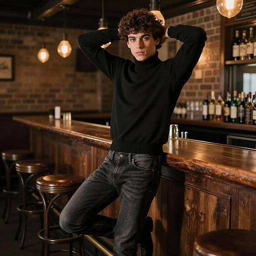 Photograph of a curly-haired young man in a black sweater and dark jeans, leaning on a wooden bar, hands behind head, in a dimly