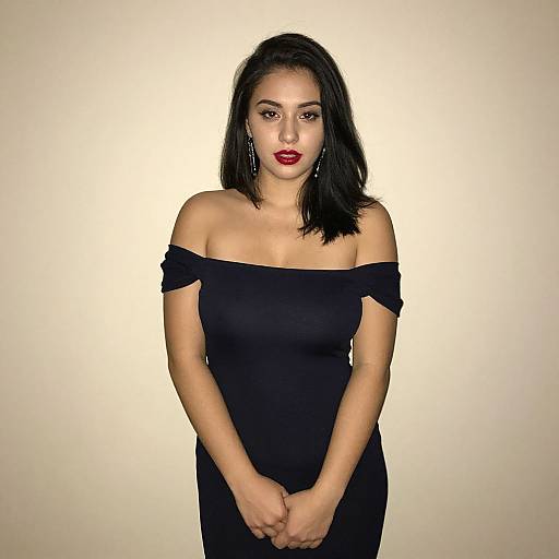Photograph of a Latina woman with long black hair, wearing an off-shoulder black dress, red lipstick, and earrings, standing against a plain
