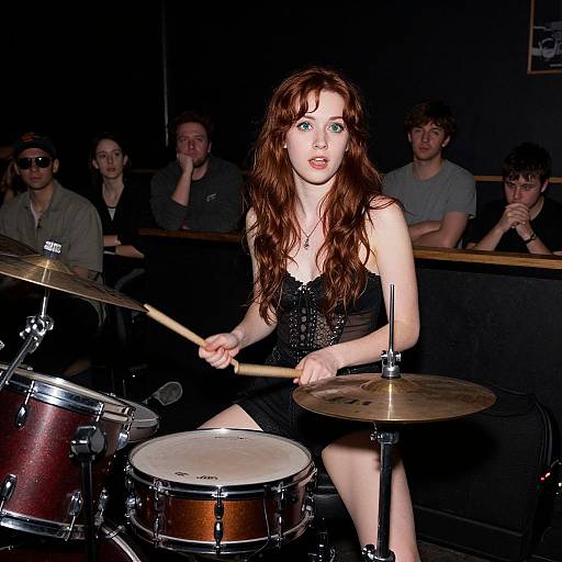 Photograph of a young, fair-skinned woman with long red hair, wearing a black sequined dress, playing drums on stage, with an audience