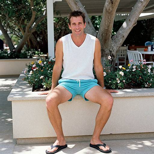 Photograph of a smiling, fit, middle-aged man with short brown hair, wearing a white sleeveless shirt, light blue shorts, and black flip