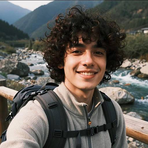 Photograph of a smiling young man with curly black hair, wearing a gray zip-up hoodie and black backpack, standing by a rocky mountain stream.
