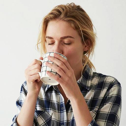 Photograph of a blonde woman with closed eyes, sipping from a white mug with black grid lines, wearing a blue and white checkered shirt,